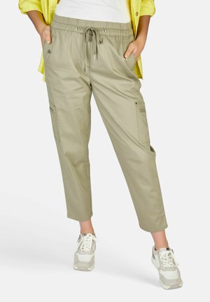 Osoba u bež hlačama s trakama za vezanje u struku i džepovima, bijelim tenisicama i žutom majicom, s rukama u džepovima.