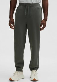 Sötétzöld, puha anyagból készült melegítőnadrág, rugalmas derekkal, zsinórral és szűkített bokákkal. Világos tornacipővel párosítva.