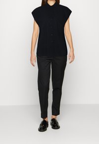Camicia nera a maniche corte con colletto strutturato, chiusura con bottoni e tessuto liscio; abbinata a pantaloni neri slim-fit e mocassini lucidi.