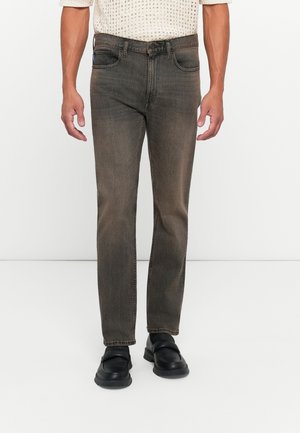 Mann iført falmede brune straight-leg jeans, sorte sko og en hvid strikket sweater som står mot en hvit bakgrunn.