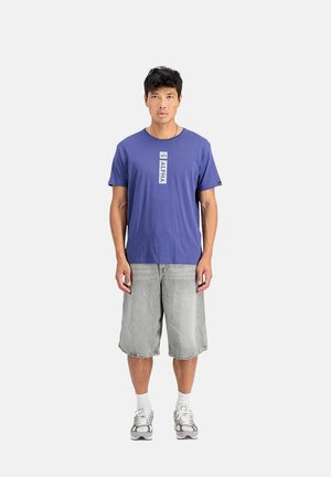 Lila Baumwoll-T-Shirt mit dem weißen Text "ALPHA", kombiniert mit knielangen, hellgrauen Jeansshorts und grauen Sportschuhen.