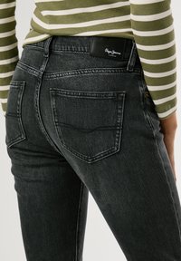 Melni džinsu bikses ar pieguļošu dizainu, kas iekļauj klasisko piecu kabatu stilu un melnu ādas etiķeti uz jostas.