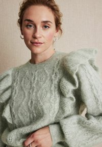 Jasnozielony sweter z dzianiny z warkoczowym wzorem i falbankowymi rękawami, wykonany z miękkiej włóczki. Modelka trzyma sweter, prezentując jego strukturę i design.