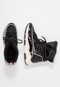 Czarne botki za kostkę z błyszczącym wykończeniem lakierowanym, cholewką z tkaniny, sztucznym futerkiem w środku oraz grubą białą podeszwą. Posiadają czarne sznurowadła i klasyczny design.