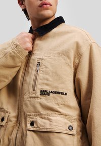 Beige jakke laget av bomull med en svart kordfløyelskrage, med en glidelåslomme og logo broderi i svart. Knapper er inkludert.
