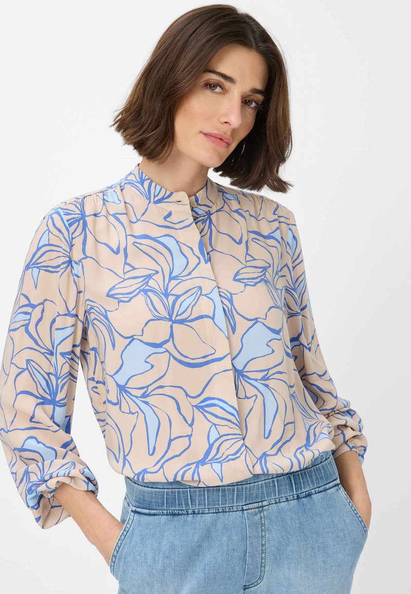 Camicia beige chiaro con motivo floreale blu; presenta un colletto alla mandarina, maniche lunghe e una vestibilità rilassata. Tessuto morbido e leggero.