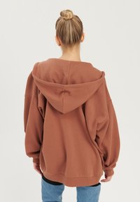 Felpa over-size marrone con un grande cappuccio, realizzata in stoffa morbida, dotata di spalle scese e polsini a costine, abbinata a jeans blu.