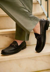 Mocassins en cuir noir à finition texturée, ornés d'un accent en métal doré. Portés avec un pantalon vert olive, sur un fond en marbre.
