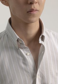 Camisa de botones en color beige claro con rayas verticales blancas. Presenta un cuello estructurado y pequeños botones, exhibiendo una textura de tela suave.