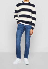 Man i marinblå och vit randig tröja, blå jeans, vita sneakers, stående med en hand i fickan mot en plain bakgrund.