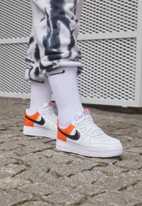 Vita Nike Air Force 1-sneakers med svart swoosh, orange detaljer och perforerad tåbox. Stylad med grått tie-dye-joggingbyxor och vita strumpor.