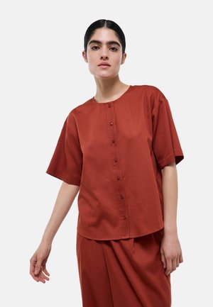 Femme aux cheveux foncés en chignon bas porte un chemisier à manches courtes couleur rouille, boutonné, et un pantalon assorti sur un fond clair uni.