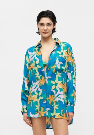 Femeie purtând o cămașă florală colorată, supradimensionată, cu top de bikini verde vizibil dedesubt, în picioare pe un fundal alb simplu.