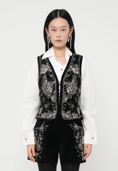 Woman with straight black hair wears white collared shirt, black floral embroidered vest, matching shorts, and long crystal drop earrings.