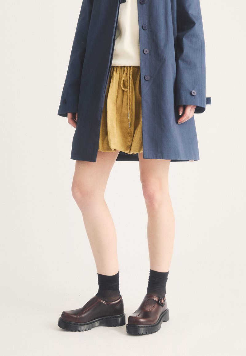 Jambes portant un manteau bleu marine, une jupe courte jaune, des chaussettes noires et des chaussures en cuir marron foncé épaisses sur un fond clair.