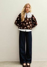 Vrouw met lang blond haar, gekleed in een sweater met luipaardprint en een grote witte kanten kraag, donkere wijdere jeans en bruine schoenen.