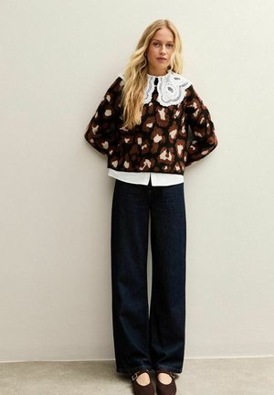 Femme aux longs cheveux blonds portant un pull en imprimé léopard avec un grand col en dentelle blanche, un pantalon large foncé et des chaussures marron.
