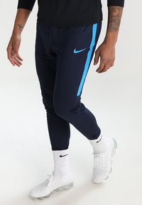 Granatowe spodnie sportowe z niebieskim paskiem bocznym, elastyczny materiał, tapered fit, zamek błyskawiczny przy mankietach, noszone z białymi butami i skarpetkami Nike.