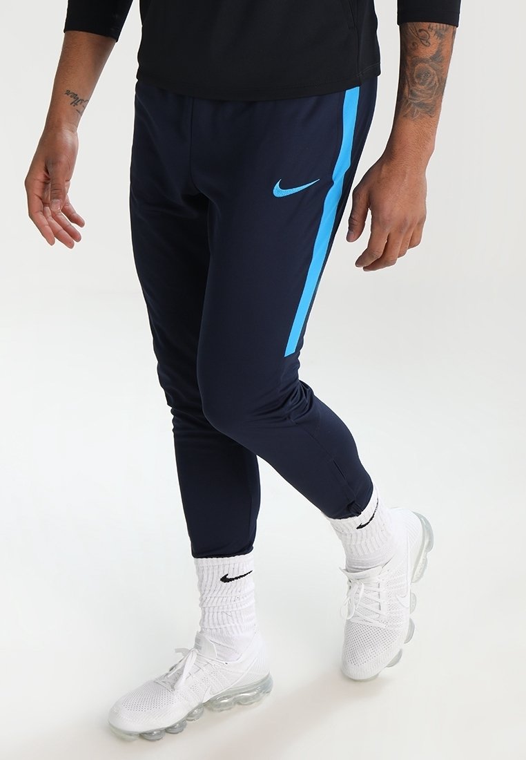 Granatowe spodnie sportowe z niebieskim paskiem bocznym, elastyczny materiał, tapered fit, zamek błyskawiczny przy mankietach, noszone z białymi butami i skarpetkami Nike.