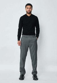 Polo noir à manches longues avec un col zippé, associé à un pantalon gris clair. Le pantalon présente un motif à carreaux subtil et une coupe ajustée.