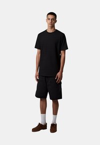T-shirt nera a maniche corte con texture, abbinata a pantaloni cargo neri. Il modello indossa scarpe marroni e calzini bianchi. Sfondo neutro.