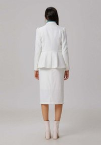 Femme se tenant de dos, portant un blazer blanc, une jupe crayon assortie, des bottines roses claires et un col roulé bleu visible.