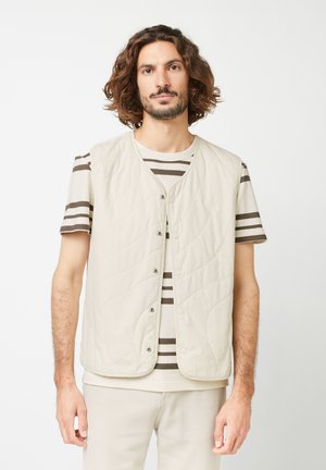 Light beige quilted vest with snap buttons, featuring a round neckline. Underneath, a striped short-sleeve shirt in neutral tones.