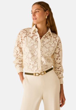 Donna che indossa una camicia crema in pizzo floreale con bottoni e maniche lunghe, pantaloni crema a vita alta, una cintura scura con fibbia dorata e orecchini a cerchio dorati.