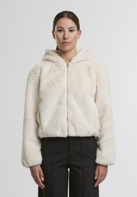 Helle, cremefarbene Kunstfelljacke mit Kapuze, Reißverschluss vorne, abgerundetem Saum und elastischen Bündchen, kombiniert mit schwarzen Hosen.