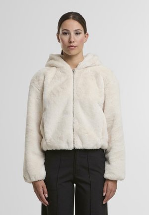 Helle, cremefarbene Kunstfelljacke mit Kapuze, Reißverschluss vorne, abgerundetem Saum und elastischen Bündchen, kombiniert mit schwarzen Hosen.