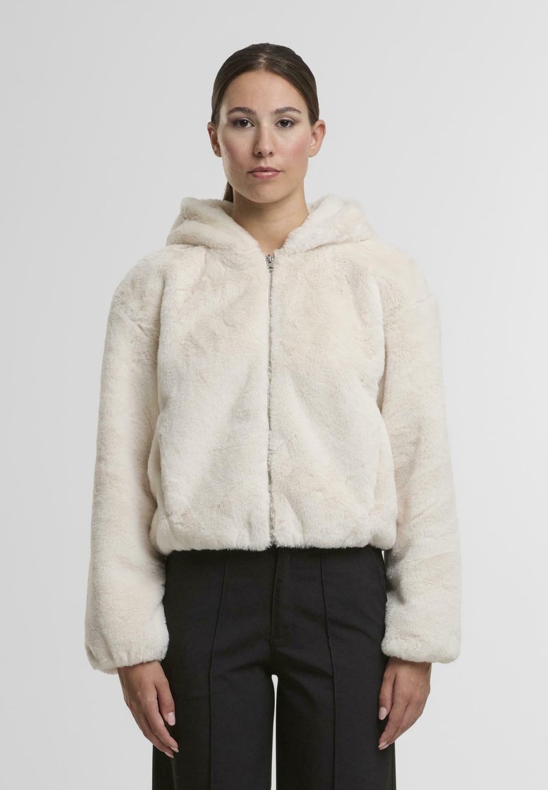 Helle, cremefarbene Kunstfelljacke mit Kapuze, Reißverschluss vorne, abgerundetem Saum und elastischen Bündchen, kombiniert mit schwarzen Hosen.
