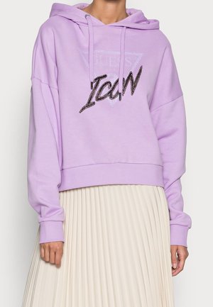 Person trägt einen hellvioletten Hoodie mit dem Text „"ICON"“ und einem schwach sichtbaren Logo „"Guess"“, kombiniert mit einem weißen Faltenrock.