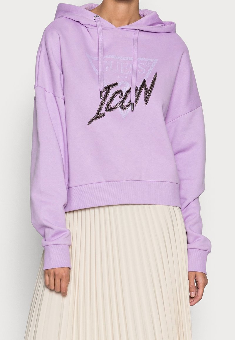 Personne portant un sweat à capuche violet clair avec le texte « "ICON" » et un logo discret « "Guess" », associé à une jupe plissée blanche.