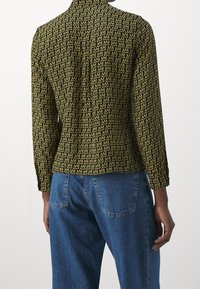 Camicia a maniche corte verde e gialla con un motivo ripetuto di lettere 'M'. Presenta un colletto a punta e polsini abbottonati. Indossata con jeans blu.
