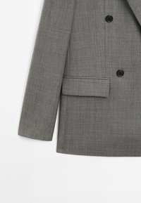 Manche et devant de blazer gris ajusté avec deux boutons noirs et une poche à rabat sur fond blanc.