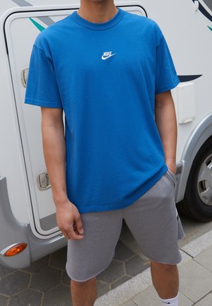 Camiseta de algodón azul con un logo blanco de Nike en el pecho, combinada con pantalones cortos grises. La textura es suave y el diseño es casual.