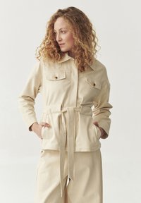 Beige puuvillatakki, jossa on vyötärölle solmittava vyö, kaksi etutaskua, nappikiinnityksellä varustetut läppäkiinnitykset ja klassinen kaulus. Sujuva pinta, rento muotoilu.