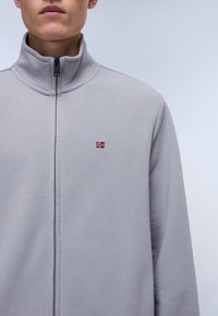 Šedá fleecová bunda na zip s vysokým límcem, která má na hrudi malou vyšívanou norskou vlajku. Jednoduchý design s jemnou texturou.