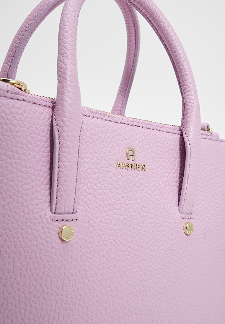 Lilafarbenes Lederhandtasche mit strukturiertem Finish, zwei Griffen, goldenen Beschlägen und einem Logodetail auf der Vorderseite. Reißverschluss oben für sicheren Verschluss.