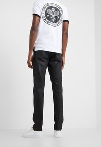 Bílé tričko s černými detaily, na zádech se nachází kruhový motiv. Černé džíny s úzkým střihem. Bílá sportovní obuv dokončuje outfit.