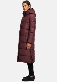 Kvinna i en lång vinröd vadderad vinterjacka med huva, svarta byxor och svarta sneakers, som står i profil mot en ljus bakgrund.