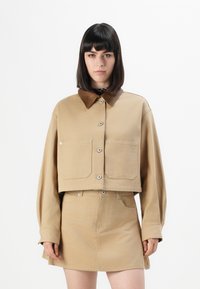 Kvinne iført en beige cropped jakke med store framlommer og et matchende beige skjørt, stående mot en enkel hvit bakgrunn.