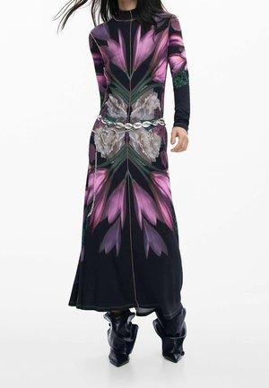 Kobieta w długiej czarnej sukience z dużymi różowo-białymi kwiatowymi wzorami, srebrnym paskiem łańcuchowym i czarnych luźnych butach w szpic.