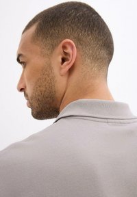 Polo grigio chiaro con un tessuto testurizzato, colletto distintivo e cuciture dettagliate. L'immagine mostra un modello maschile di profilo e di spalle.