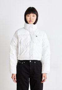 Vit puffjacka med hög krage och vadderad design. Har en liten svart logotyp på bröstet. Bärs över svarta jeans.