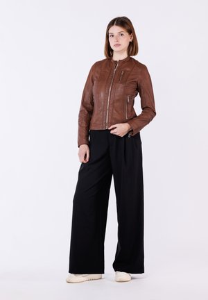 Vrouw staand, gekleed in een bruine leren jas met rits, zwarte wijde broekspijpen en witte sneakers tegen een effen witte achtergrond.