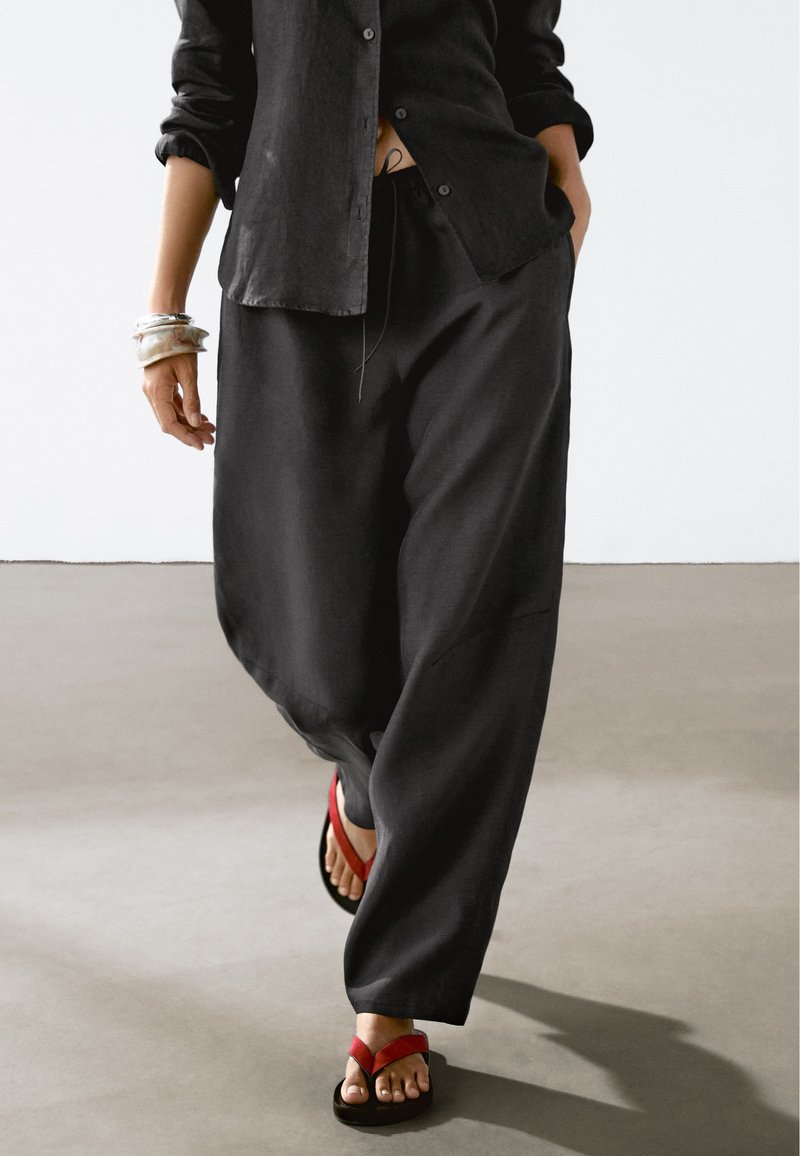 Schwarzes Leinenhemd mit Knöpfen, lockere schwarze Hosen und rot-schwarze Flip-Flops. Zubehör umfasst silberne Armbänder an einem Handgelenk.
