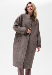 Bruine visgraatjas met hoge kraag, oversized pasvorm en zijzakken. Kenmerkt zich door een gestructureerde stof en contrasterende donkere accentkappen.