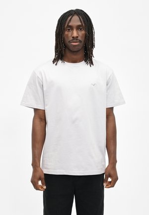 Man with dreadlocks wears plain white t-shirt and black pants, standing against a white background.