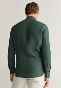 Camiseta de manga larga verde oscuro hecha de tela texturizada. Cuenta con un cuello clásico, puños con botones y un dobladillo curvado en la parte trasera.